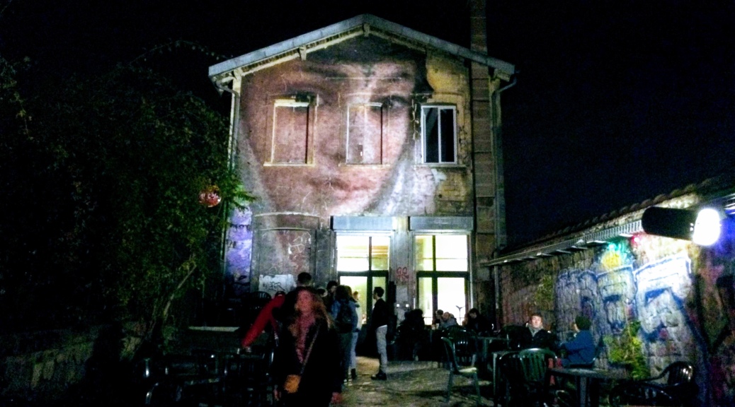 View of building facade at night and busy courtyard. Facade is painted with a large face